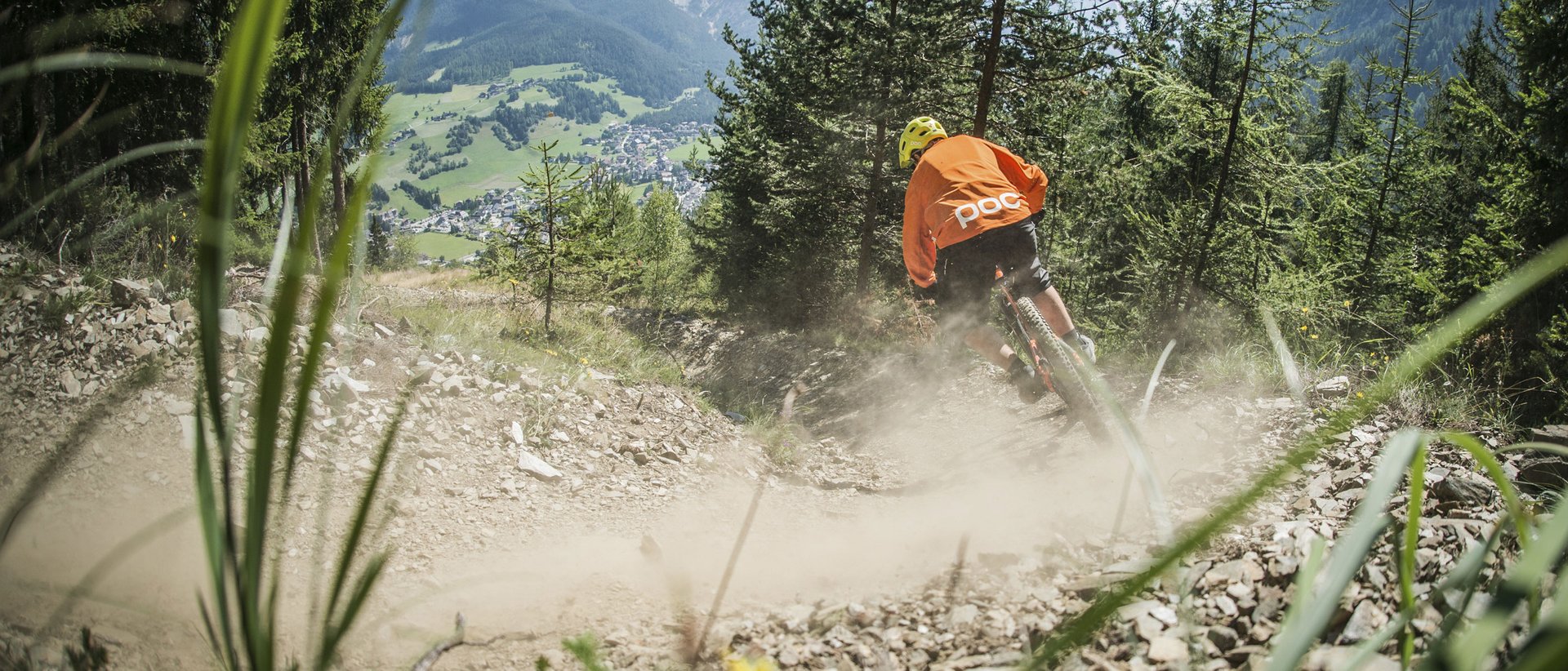 Vacanza a Plan de Corones: un’avventura Mountain biker su sentiero polveroso in montagna con boschi e villaggio sullo sfondo
