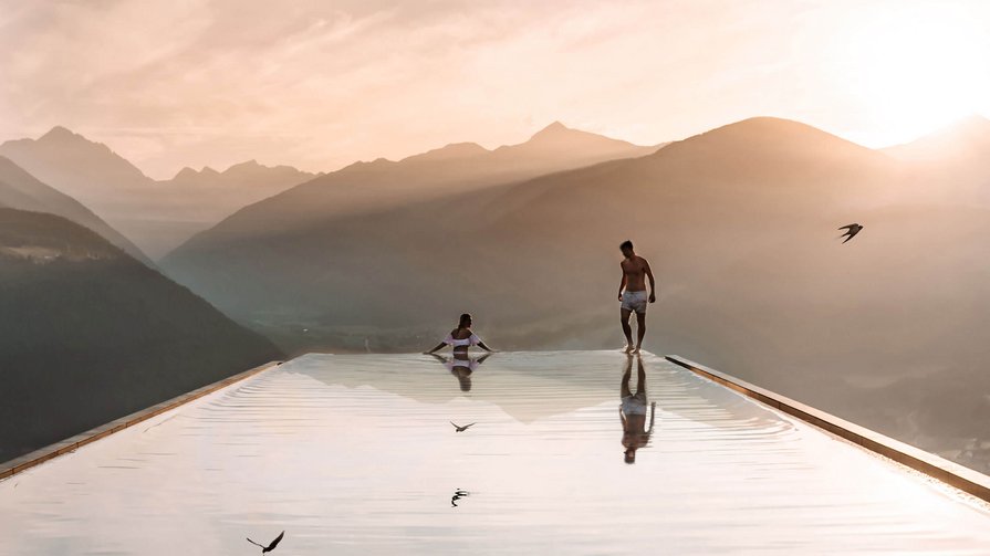 Esplorate le molteplici sfaccettature della vita nel nostro hotel e nei suoi dintorni Due persone in una piscina a sfioro con vista sulle montagne al tramonto