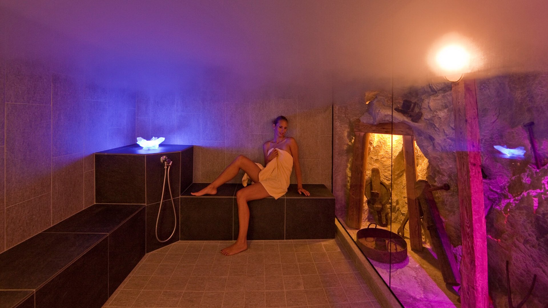 Our sauna, Plan de Corones/Kronplatz in full view Woman in towel sitting in lit steam room with stone wall and steam