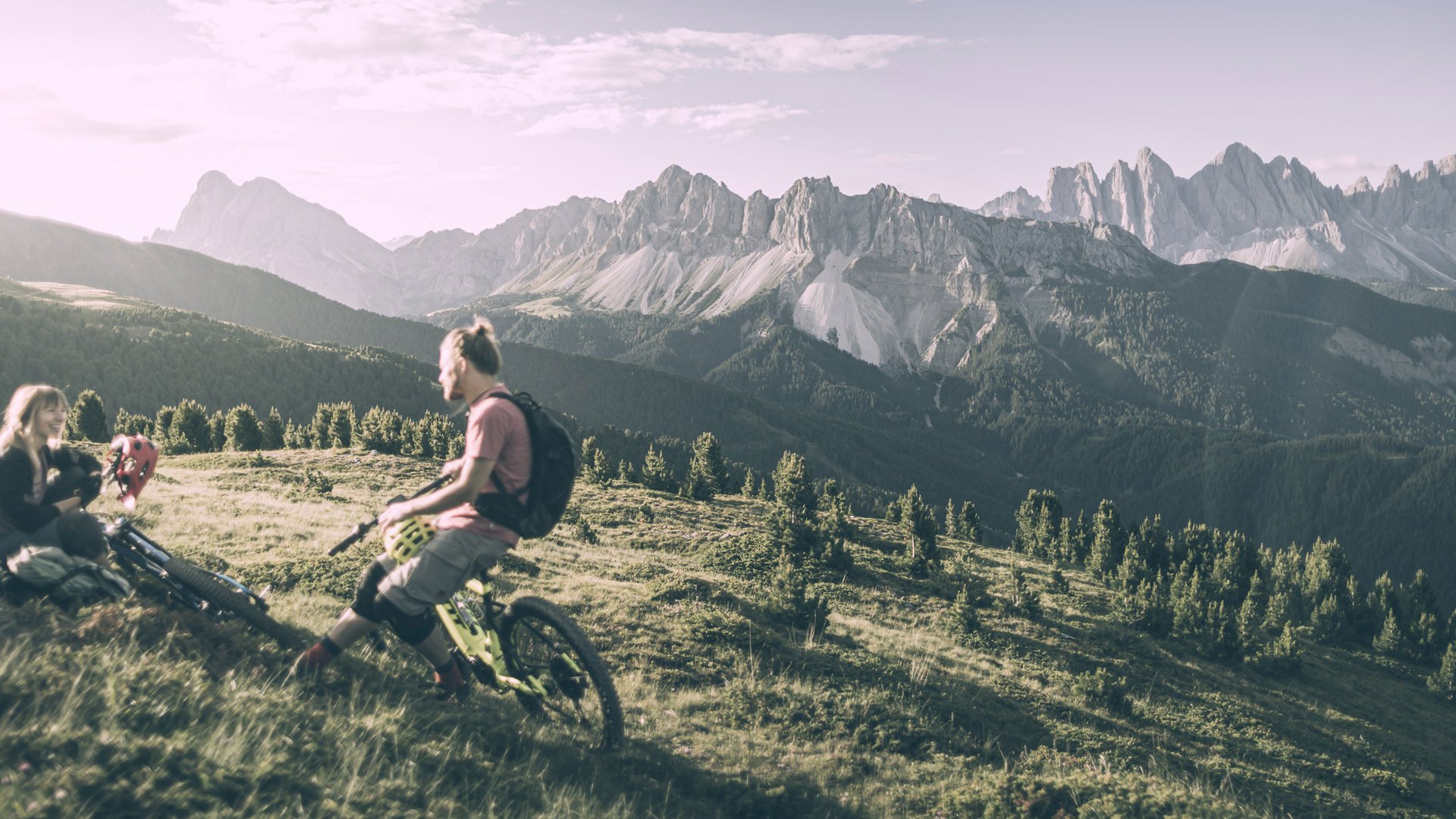 Vacanza a Plan de Corones: un’avventura Due mountain biker si riposano su un prato con panorama montano alle spalle
