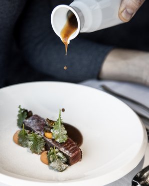 Experience moments of culinary magic Sauce being poured over a finely garnished piece of meat on a white plate