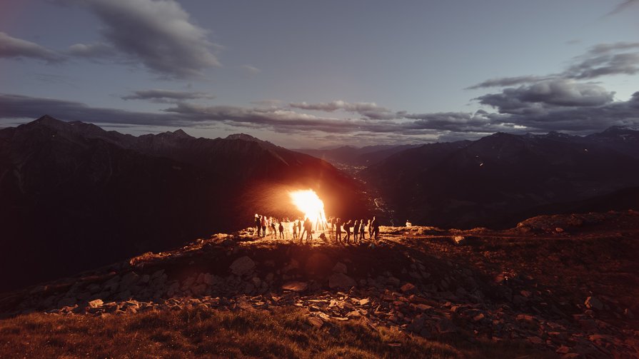 Bergfeuer – ein leuchtender Brauch Menschen um ein großes Lagerfeuer auf einem Berggipfel bei Sonnenuntergang