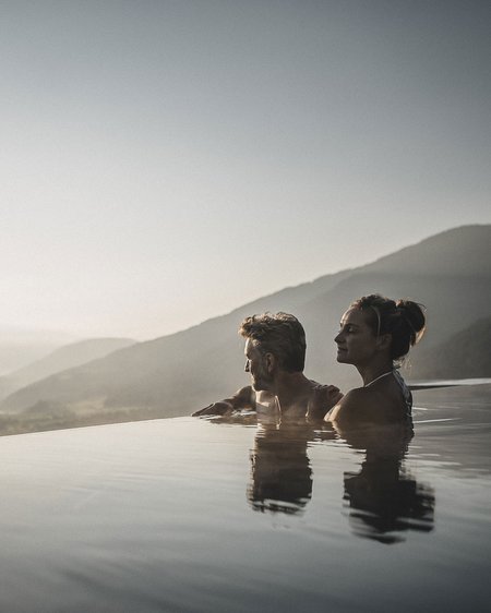 Il nostro hotel a Valdaora tra cielo e terra Coppia rilassata in piscina a sfioro con vista sulle montagne al tramonto