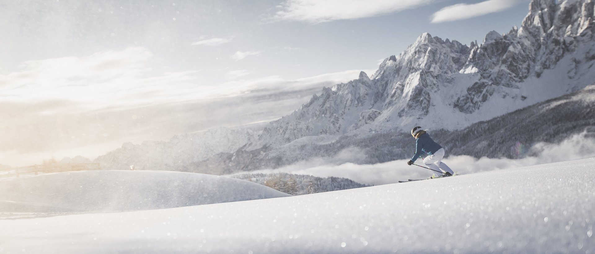 The best hotel at Plan de Corones/Kronplatz Skier descending snowy slope with rugged mountains and sunlight in background