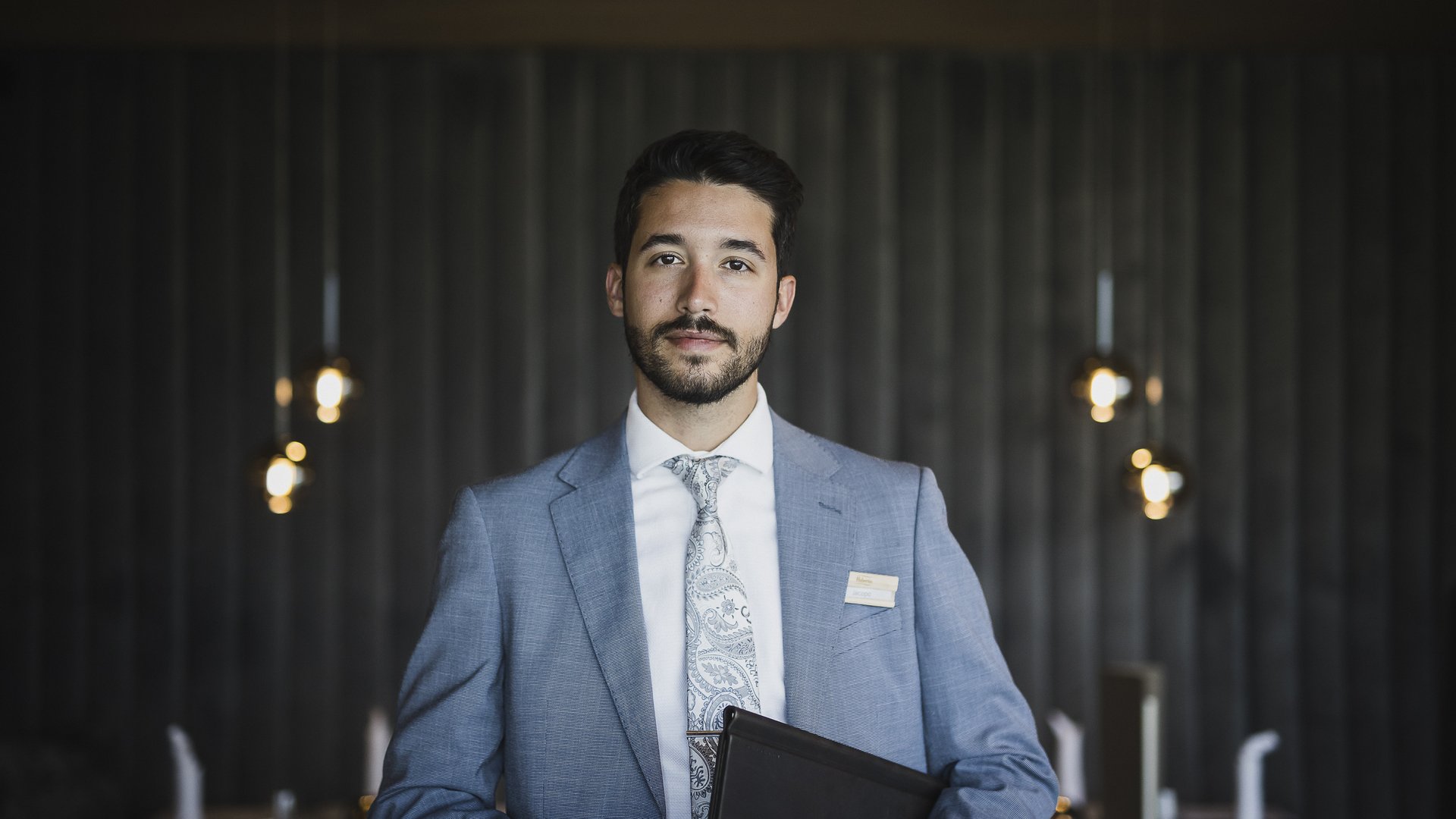 A look at our wine list Man in gray suit with tie holding black folder in modern room