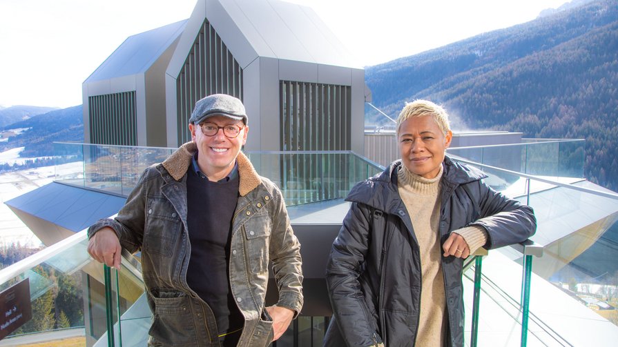 Una panoramica dell'Alpin Panorama Hotel Hubertus Due persone appoggiate a una ringhiera in vetro davanti a un edificio moderno in montagna