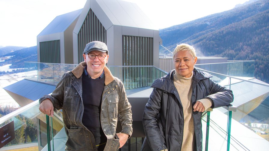 Ein Blick hinter die Kulissen des Alpin Panorama Hotels Hubertus Zwei Personen lehnen an Glasgeländer vor moderner Bergarchitektur