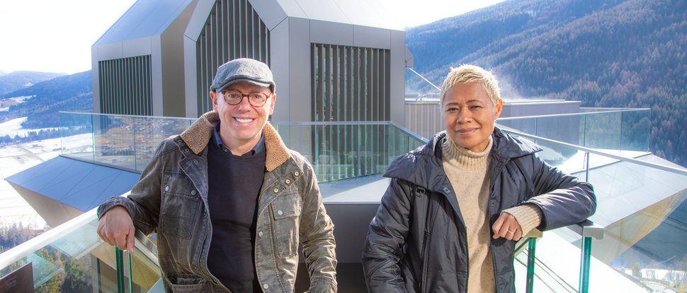 Ultime novità e storie da Valdaora Due persone appoggiate a una ringhiera in vetro davanti a un edificio moderno in montagna