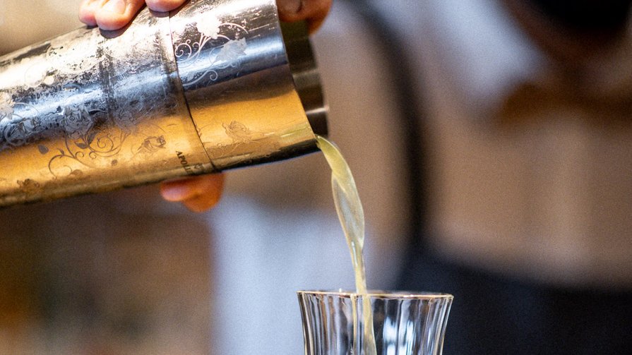 London Dry Gin | succo di limone appena spremuto | sciroppo di basilico e cetriolo | spuma di mango Un barista versa una bevanda verde da uno shaker in un bicchiere di cristallo