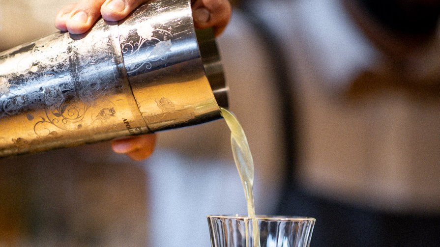 London Dry Gin | Frischer Zitronensaft | Basilikum-Gurkensirup | Mangoschaum Ein Barkeeper gießt ein grünes Getränk aus einem Shaker in ein Kristallglas