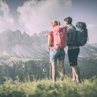 Bruneck: Shopping und mehr Wanderer mit Rucksäcken blicken auf bewaldete Berge unter bewölktem Himmel