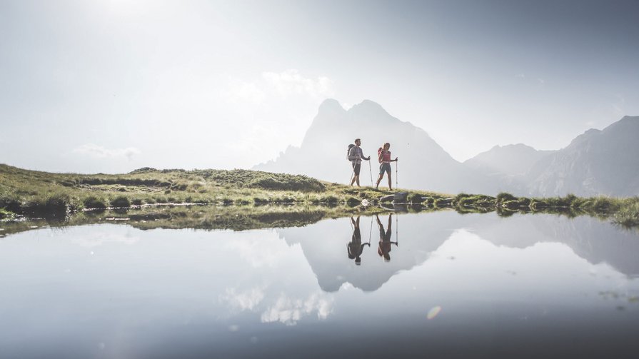Frühlingsschmelze und die Rückkehr des Lebens Zwei Wanderer mit Rucksäcken neben einem Bergsee mit Bergreflexion